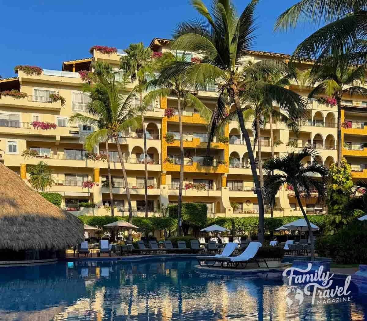 View of Velas Vallarta resort with palm trees, colorful flowers on balconies, and a large pool reflecting the yellow building under a bright blue sky.