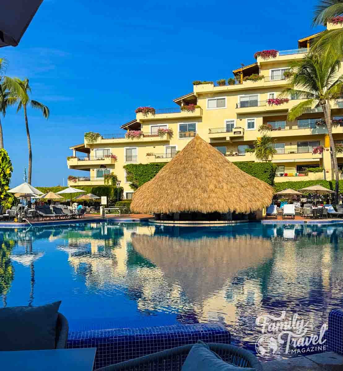 Thatched-roof pool bar surrounded by lounge chairs and palm trees at Velas Vallarta resort, reflecting in the calm blue water.
