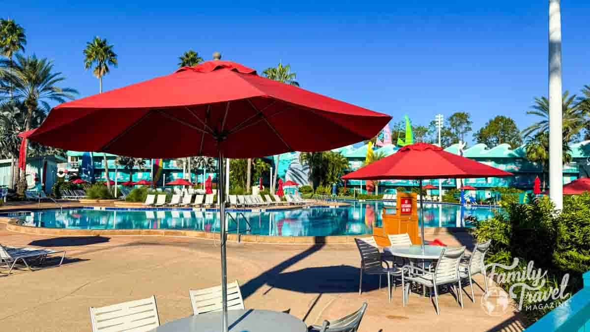 Red umbrellas and tables surround a swimming pool at a resort on a sunny day.