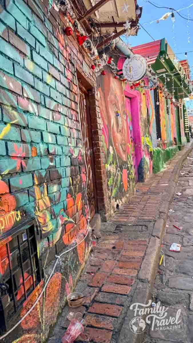 Colorful brick walls with vibrant murals line a narrow, decorated street in Bogot&aacute;, Colombia.