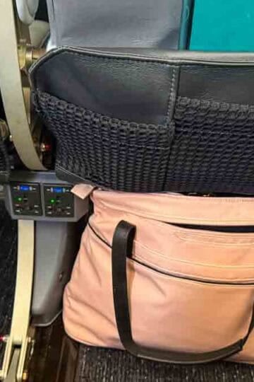 A pink travel bag is stowed under an airplane seat, with power outlets and seatback pockets visible.