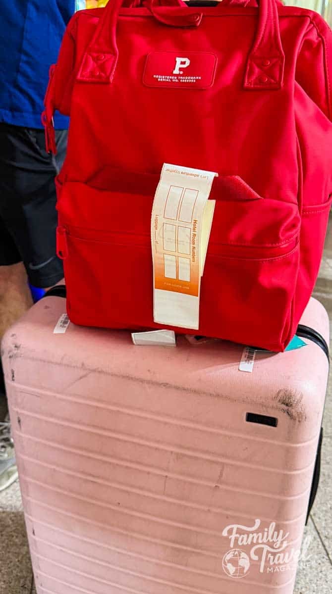 A red backpack with a luggage tag rests on a scuffed pink suitcase, ready for travel.