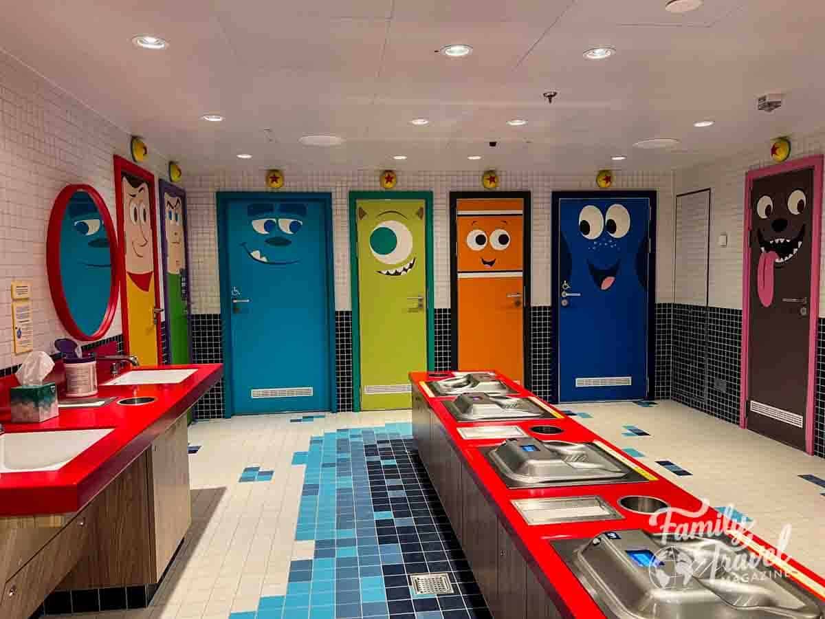 Colorful Disney Wish kids club bathroom with character-themed doors and red sinks.