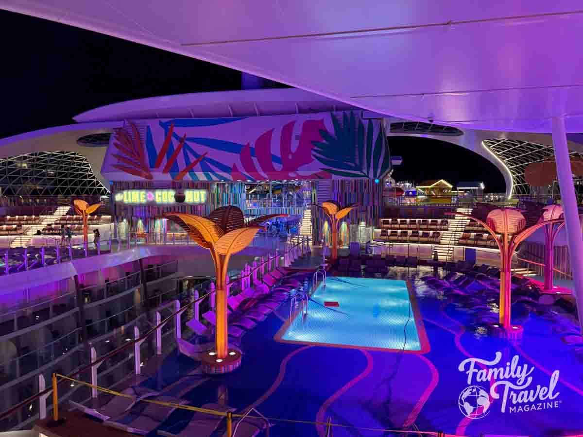The vibrant pool deck of the Disney Wish cruise ship glows with purple and pink lights at night.