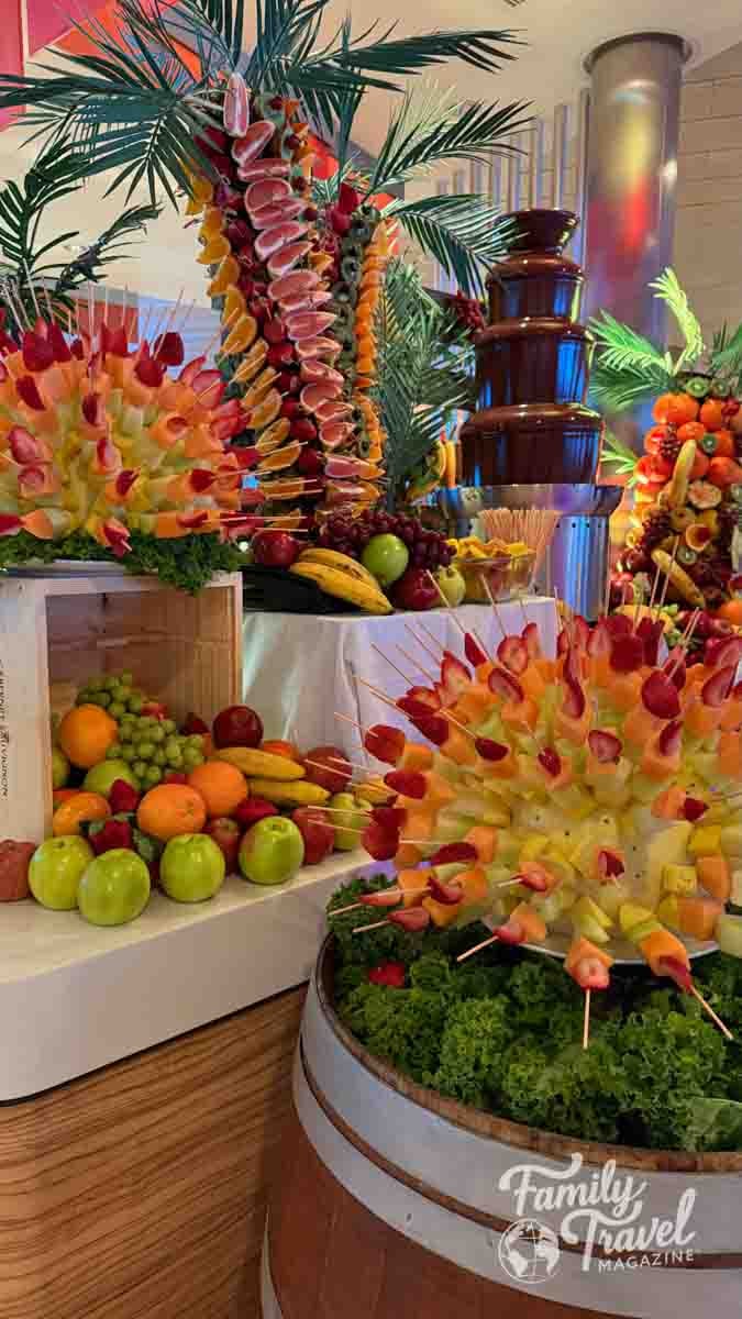 A lavish fruit and chocolate fountain buffet with palm frond decorations and colorful fruit skewers.