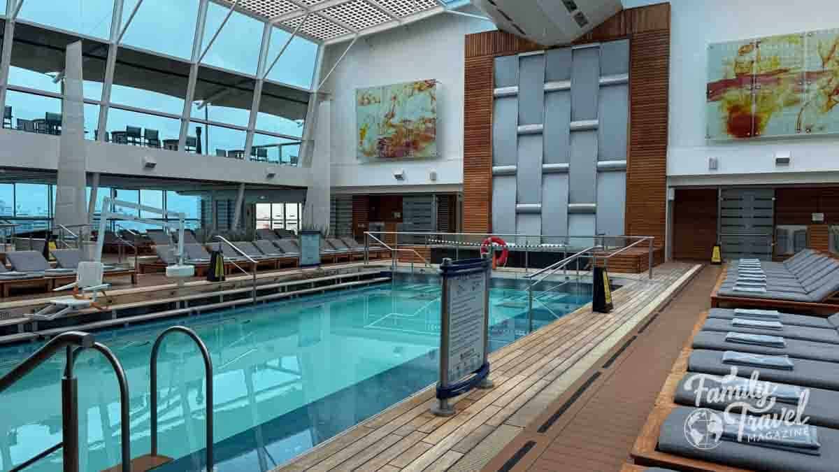 A bright, modern cruise ship solarium with an indoor pool, lounge chairs, and a large glass ceiling.