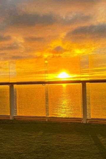 Golden sunset reflects intensely on the ocean, viewed from a cruise ship deck with green turf and glass railing.