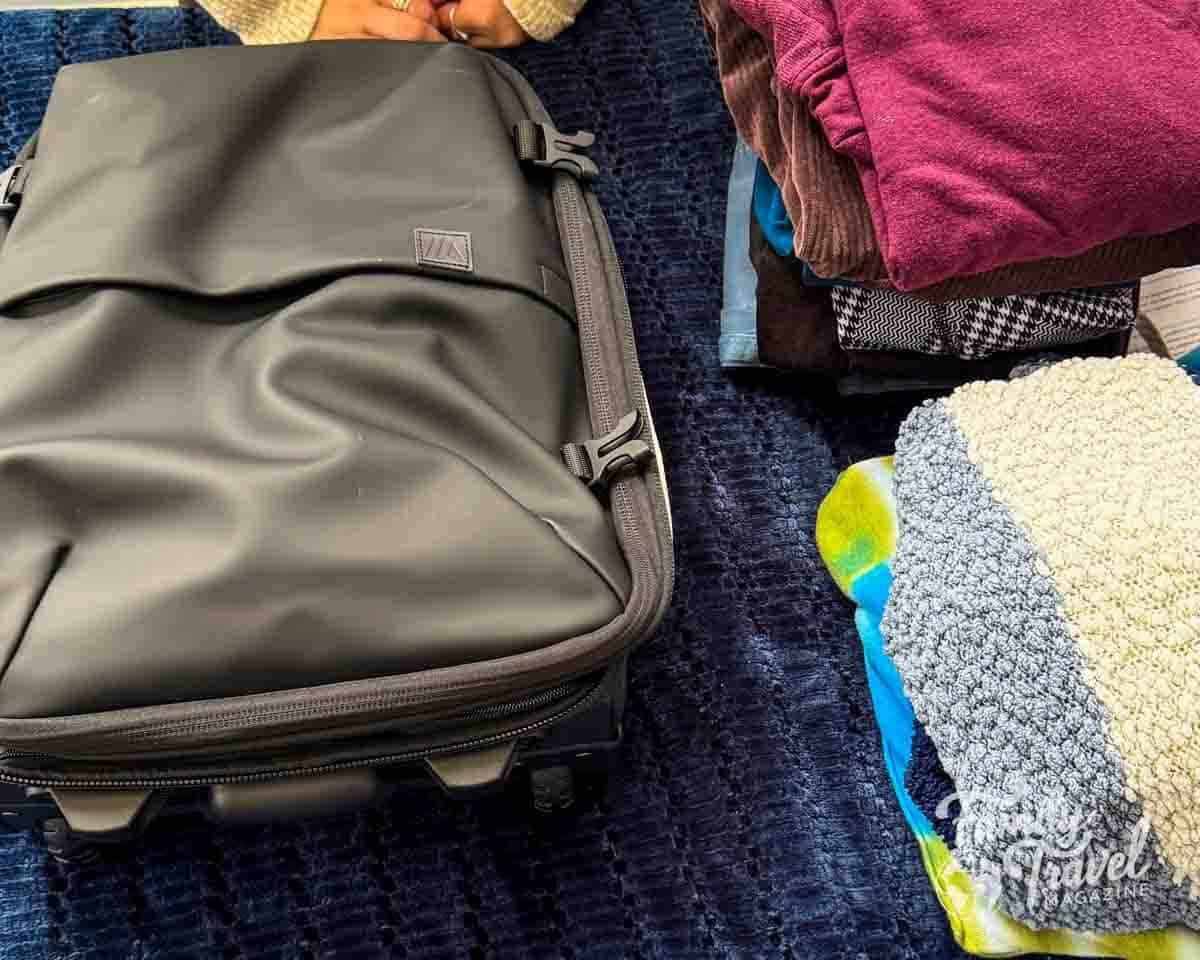 A black rolling carry-on backpack and folded clothes on a blue textured blanket.