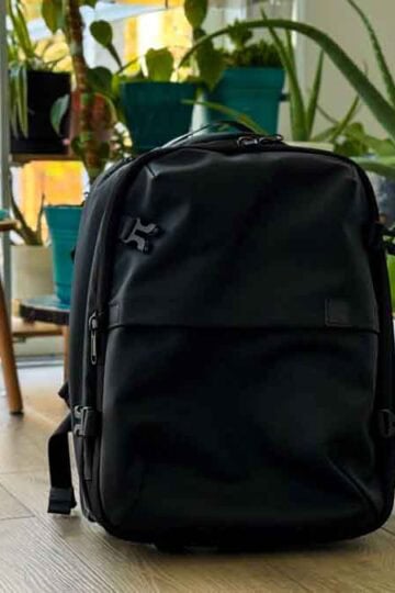 Black backpack on a light wood floor, surrounded by potted plants and a white door.