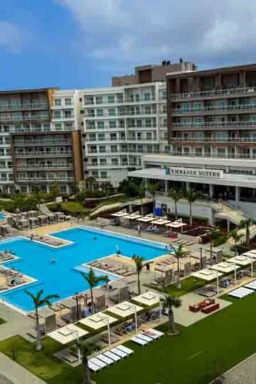 Aerial view of the Embassy Suites hotel in Aruba, featuring multiple pools, lounge chairs, and palm trees.