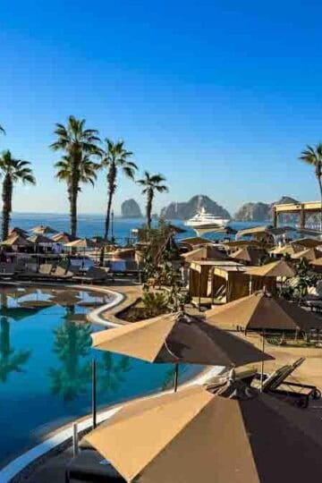 Sunny resort pool with palm trees, lounge chairs, and El Arco de Cabo San Lucas visible in the ocean.