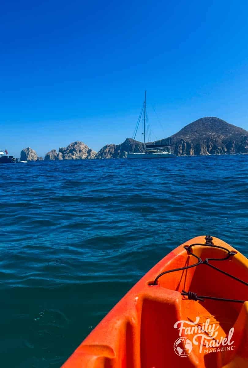 Kayaking towards El Arco de Cabo San Lucas, with a sailboat and rocky islands under a clear blue sky.