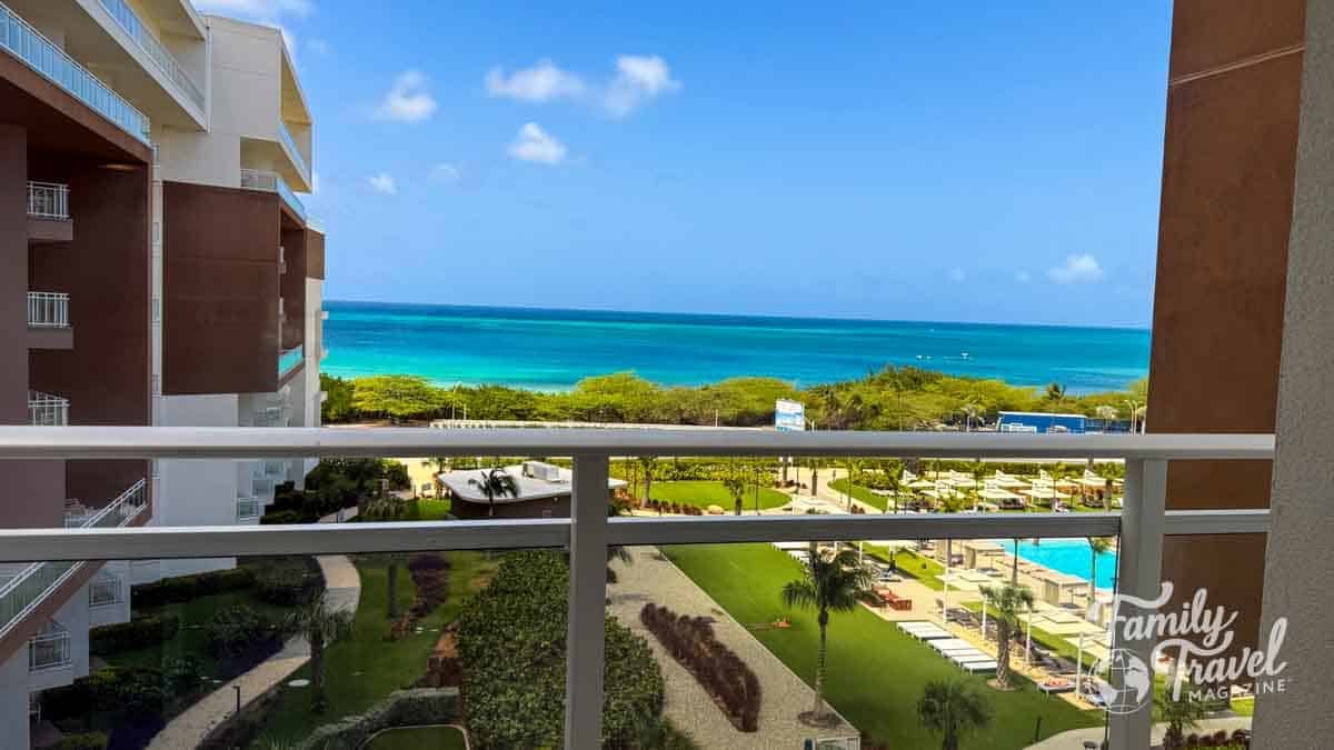 Balcony view of a resort pool, lush gardens, and the turquoise Caribbean Sea in Aruba.