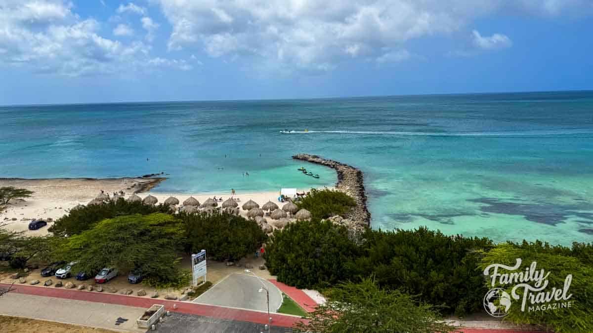 Aerial view of a beautiful beach in Aruba with turquoise water, a rock jetty, and straw palapas.
