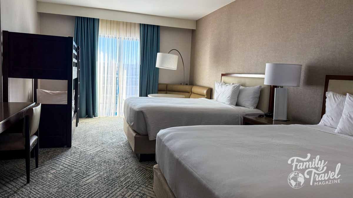 A spacious hotel room featuring two queen beds, a bunk bed, and a sofa near a window with blue curtains.