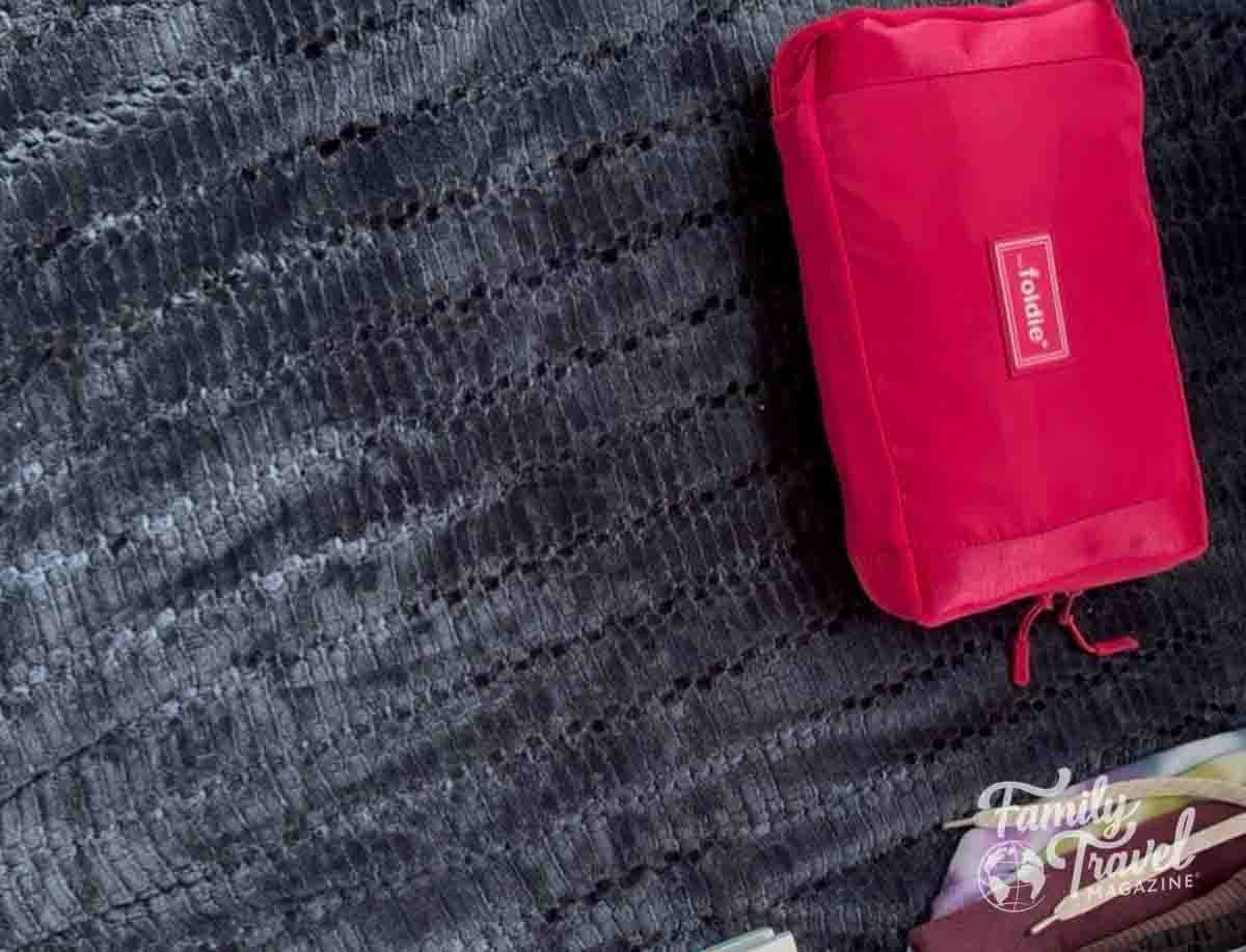 A pink travel pouch lies on a dark textured blanket, with a Family Travel Magazine logo.