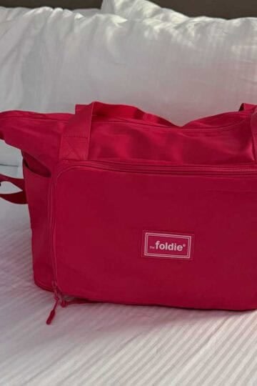 A vibrant red Foldie travel bag with a water bottle rests on a white hotel bed.