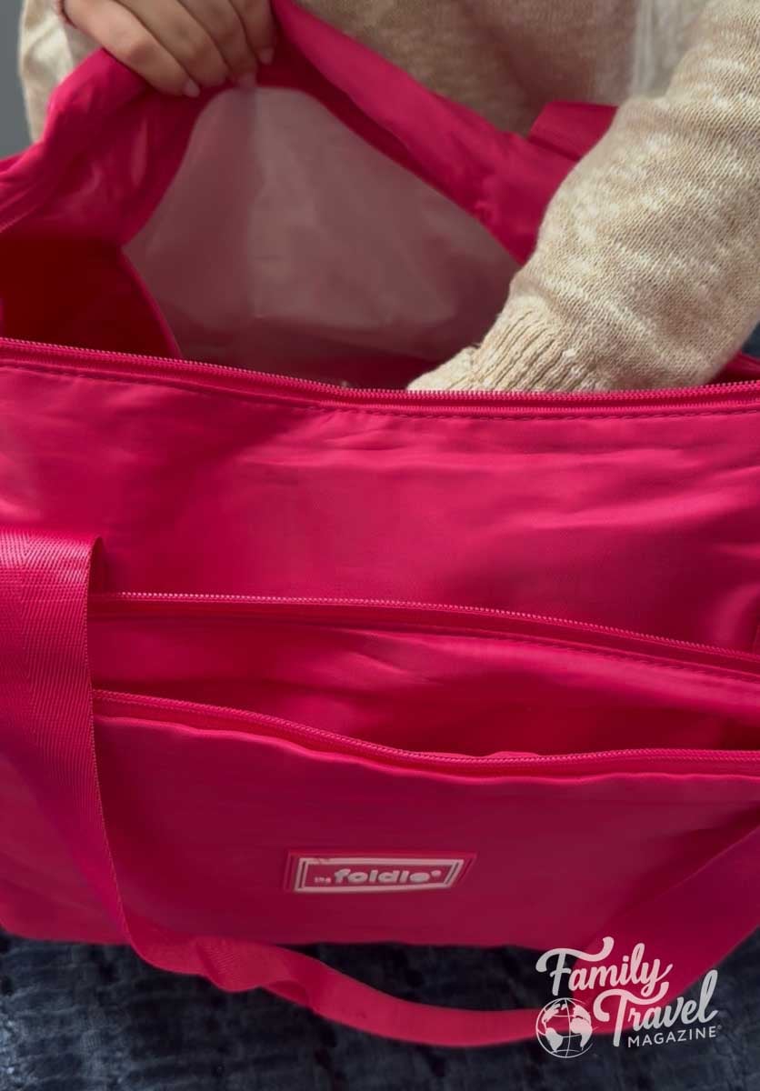A persons hand reaches into a bright pink the foldie travel bag with multiple zipper compartments.