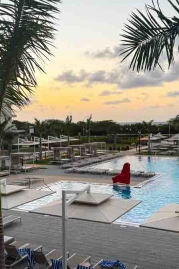 Sunset over a large resort pool area with palm trees, many lounge chairs, cabanas, and a hotel building.