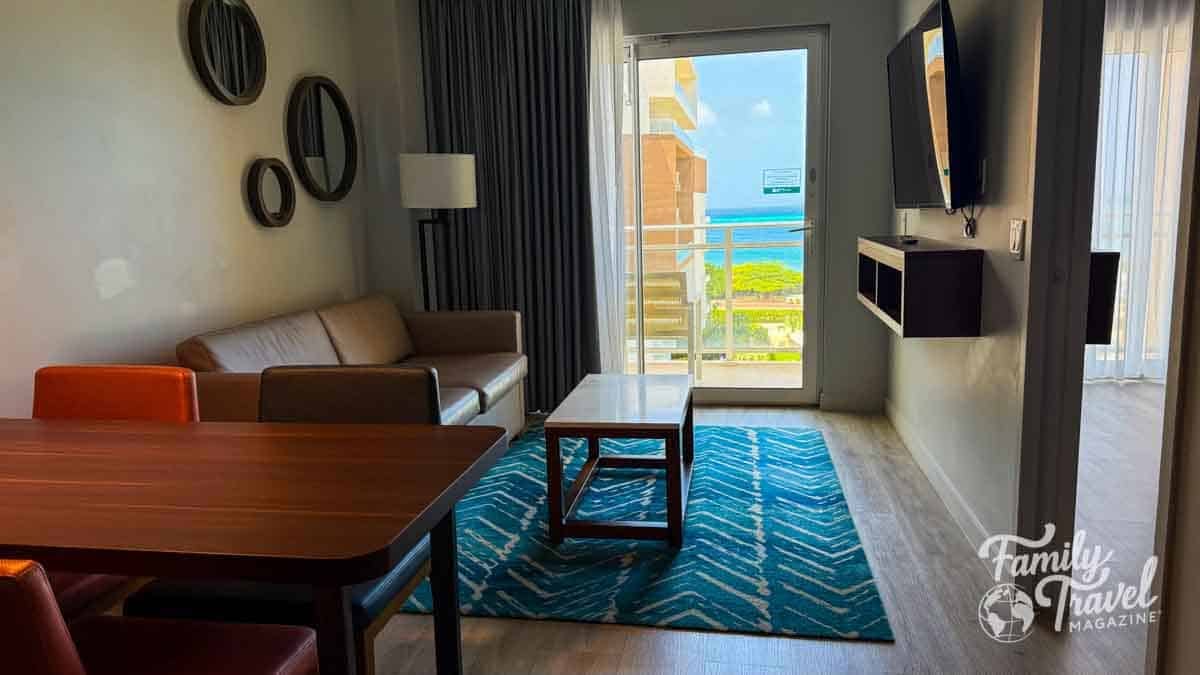 A modern resort living room with a sofa, dining table, and a balcony overlooking the turquoise ocean.