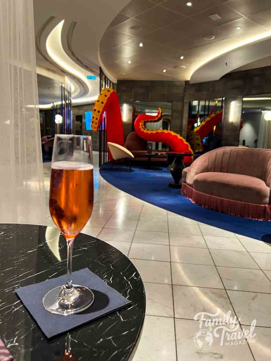 Glass of sparkling wine on a table in a stylish Virgin Voyages lounge, with modern seating and playful octopus-inspired d&eacute;cor in the background.