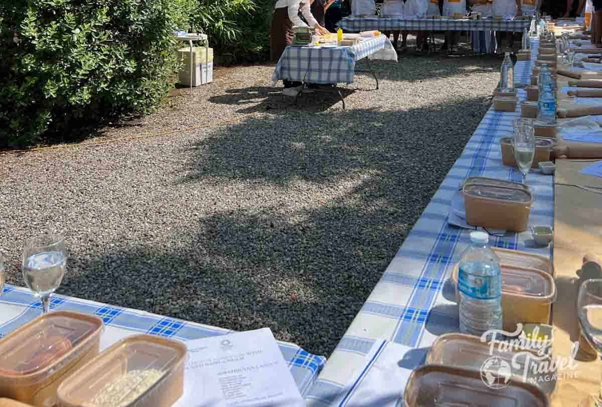 Outdoor family cooking class with individual ingredient kits and rolling pins on checkered tables.