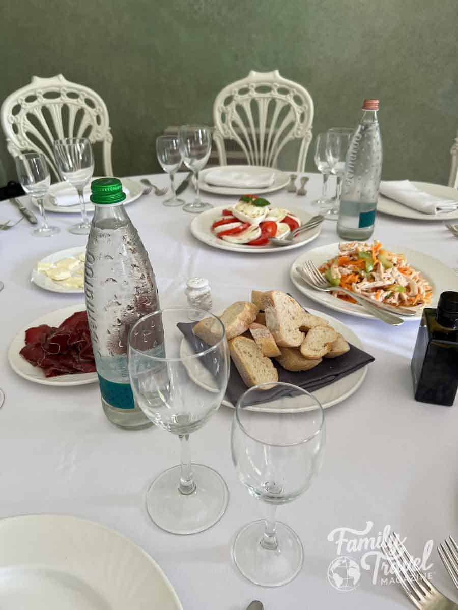 An elegant dining table set with Caprese salad, cured meat, bread, water, and ornate white chairs.