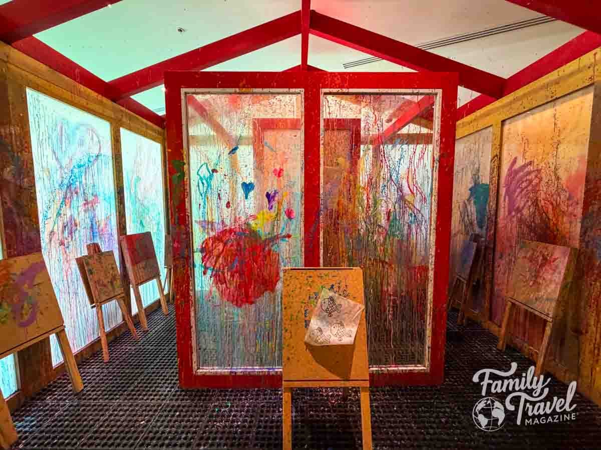 A vibrant childrens art studio with red beams, paint-splattered walls, and easels for creative family fun.