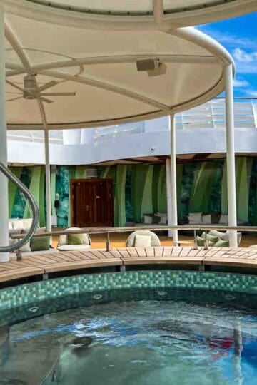 A vibrant cruise ship deck features a tiled hot tub, shaded lounge seating, and green tropical-themed walls.