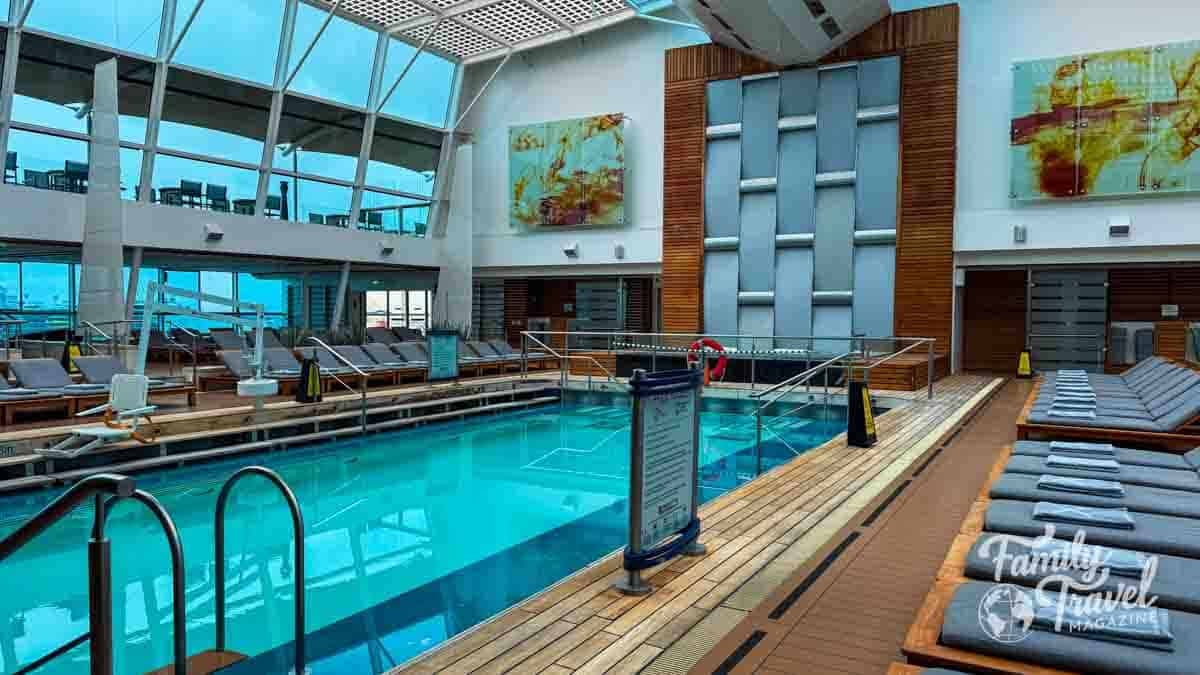Indoor solarium pool on a Celebrity Cruises ship, featuring a glass roof, heated pool, loungers, and panoramic ocean views.