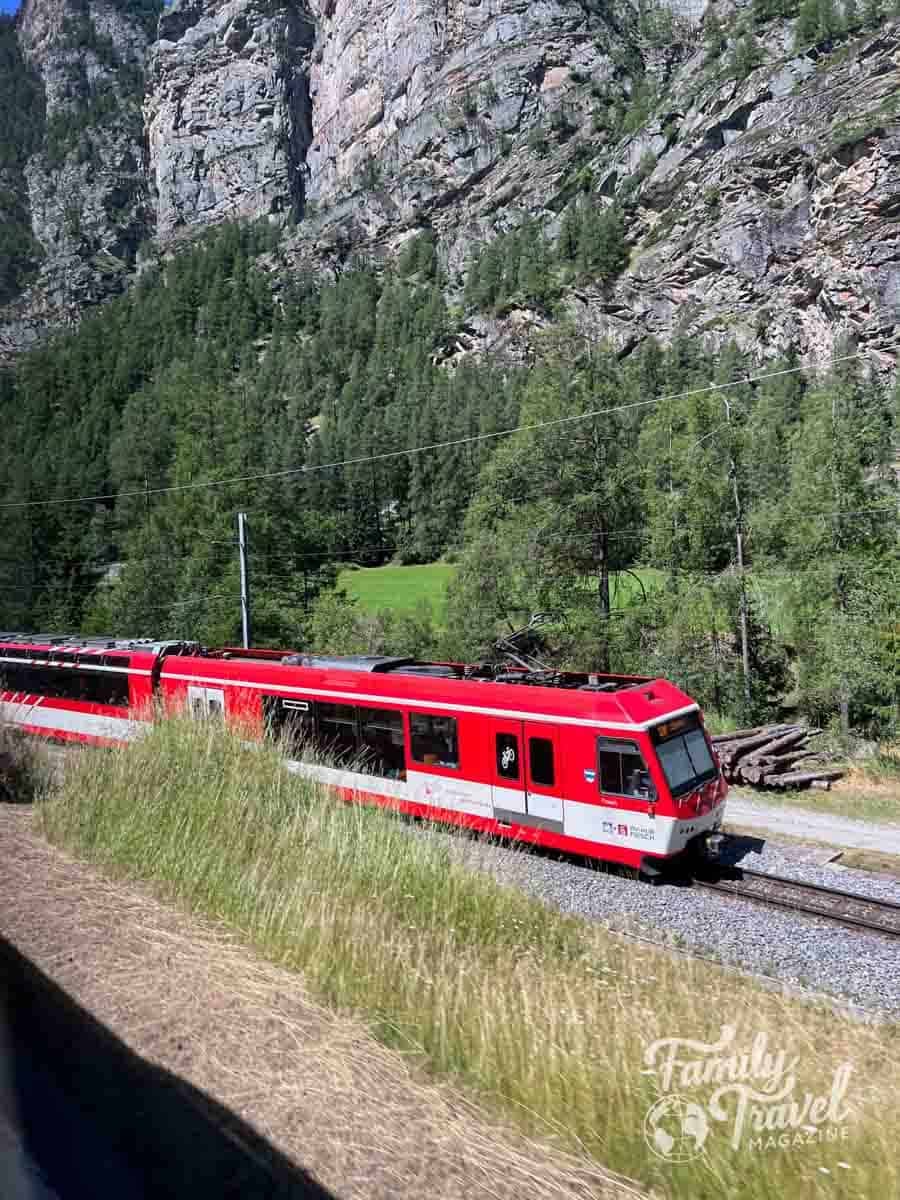 A red Rhaetian Railway train travels through the steep, forested Swiss Alps.