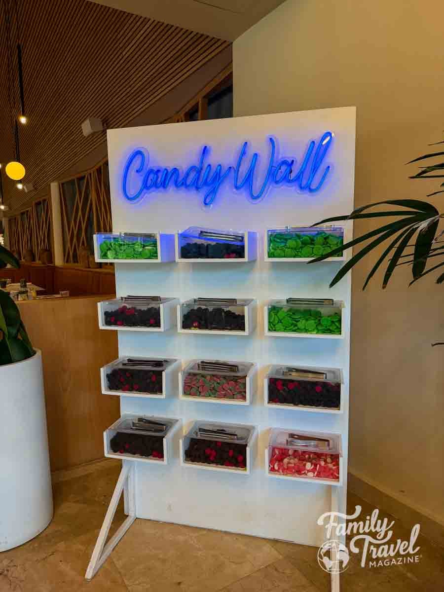 A bright blue neon Candy Wall sign illuminates a display of various colorful candies in clear bins.