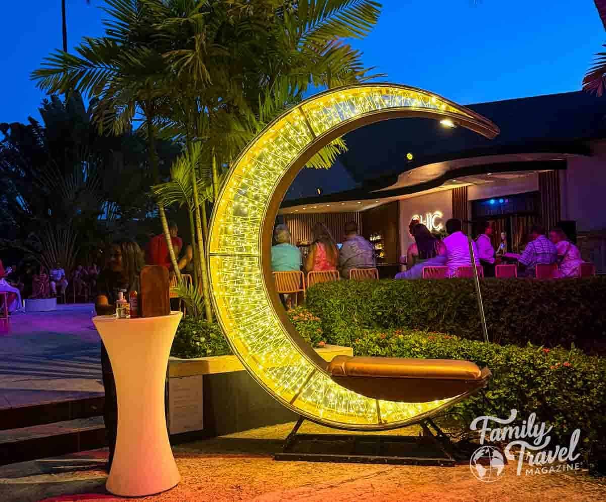 Guests relax at the illuminated CHIC by Royalton resort bar under palm trees at night.