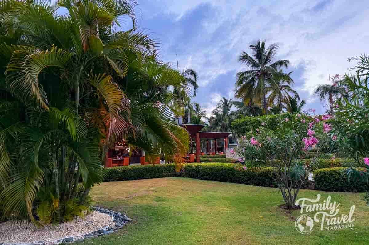 Lush tropical resort garden with palm trees, pink flowers, and an outdoor restaurant under a cloudy sky.