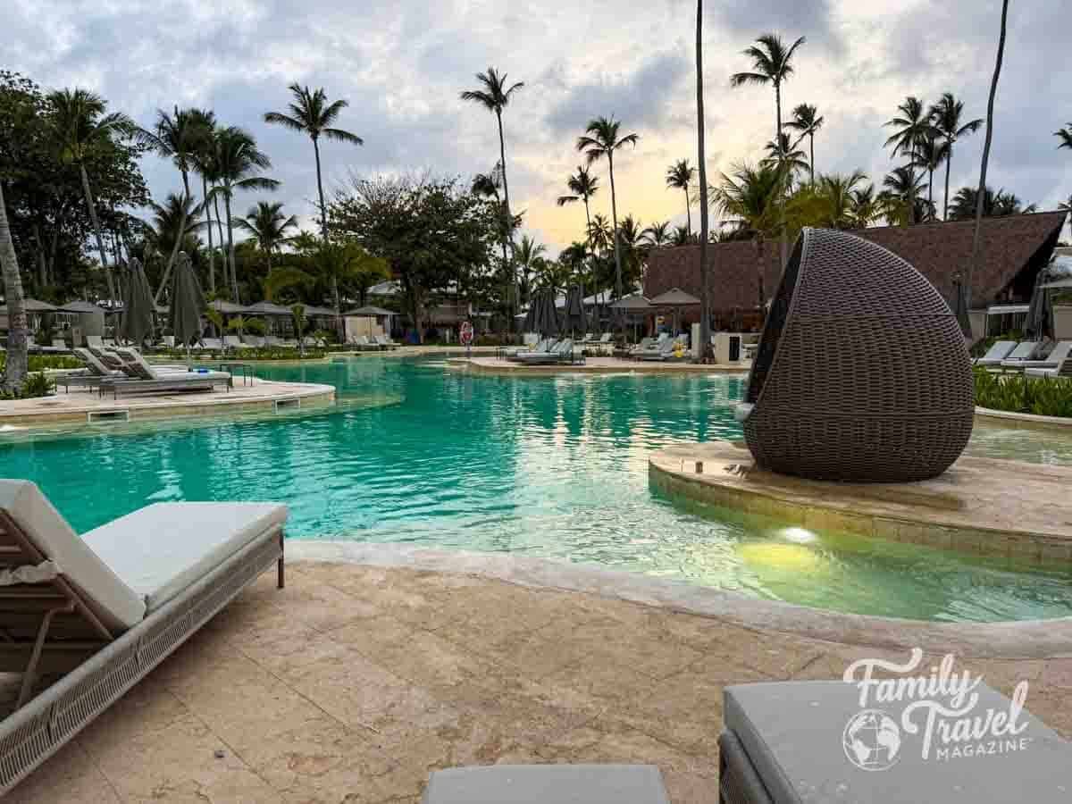 A luxurious tropical resort pool with palm trees, numerous lounge chairs, and a distinctive woven pod chair at dusk.