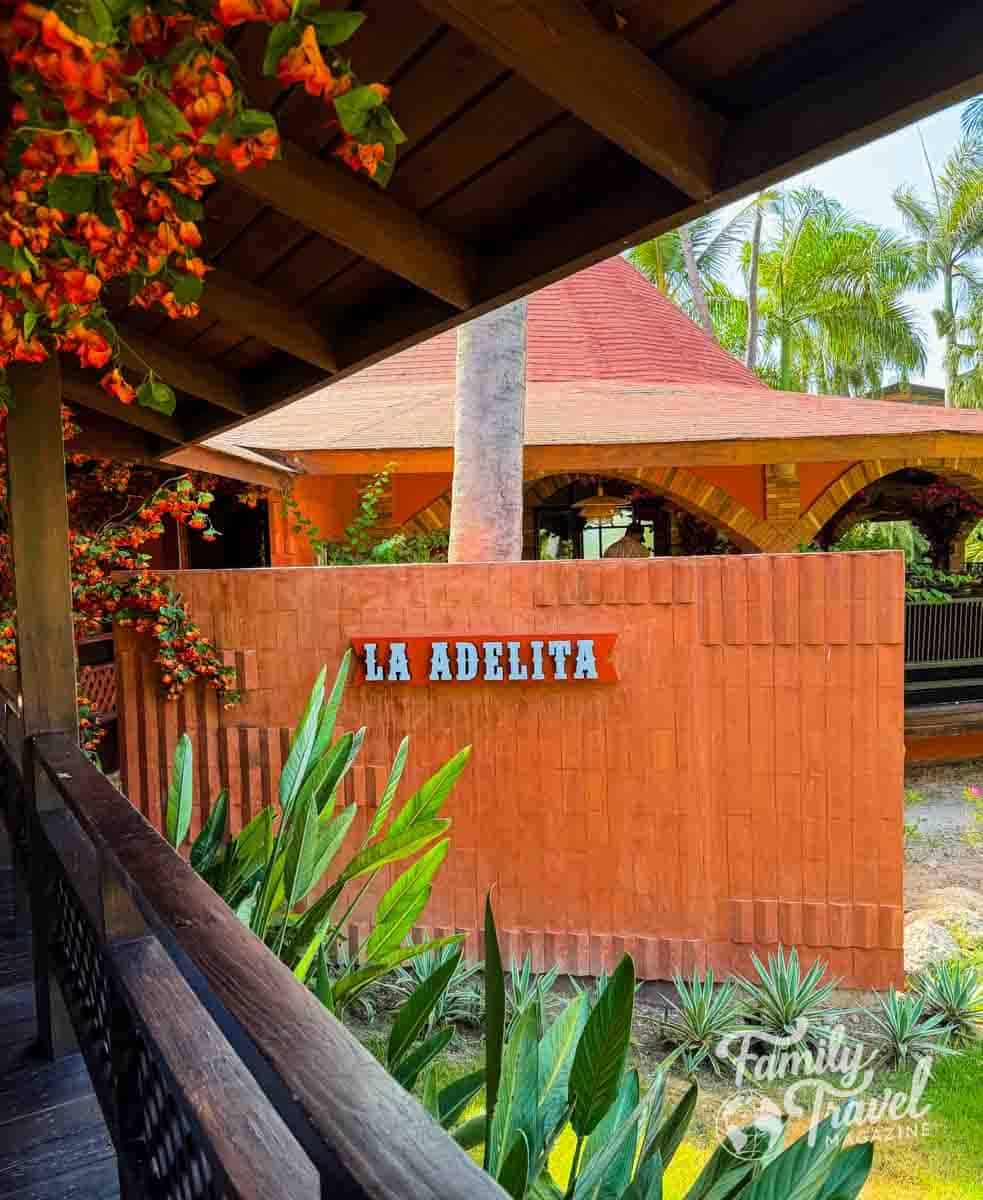 Sunny view of La Adelita restaurant with orange walls, red roof, palm trees, and vibrant orange flowers.