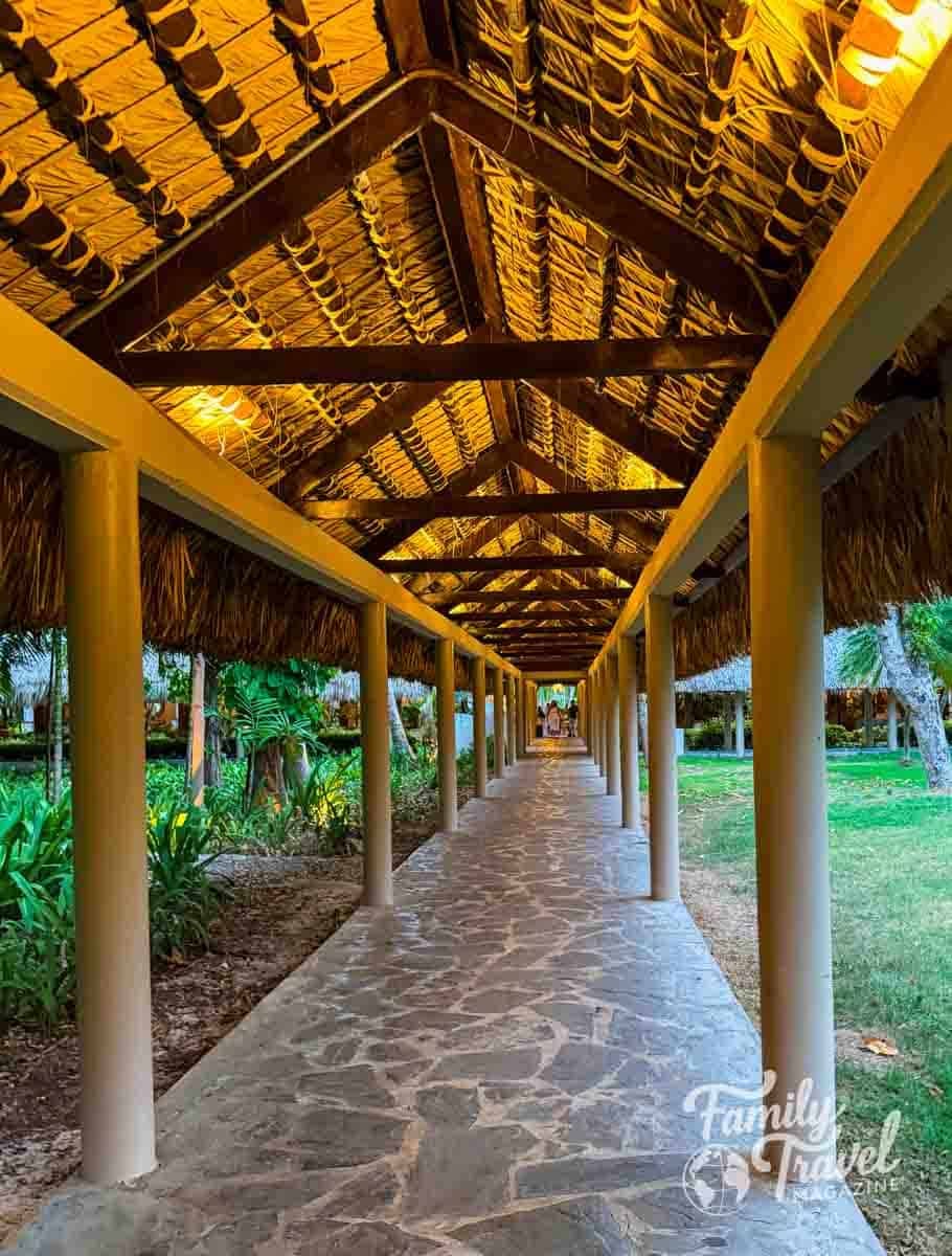 Long, illuminated thatched-roof walkway with a stone path through tropical gardens at a resort.