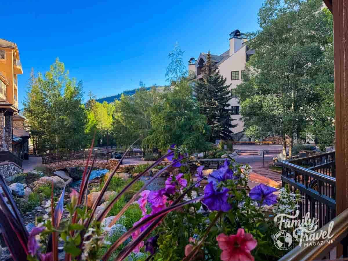 Vibrant flowers frame a view of Beaver Creek Village with a stream, stone bridge, and mountain lodges.