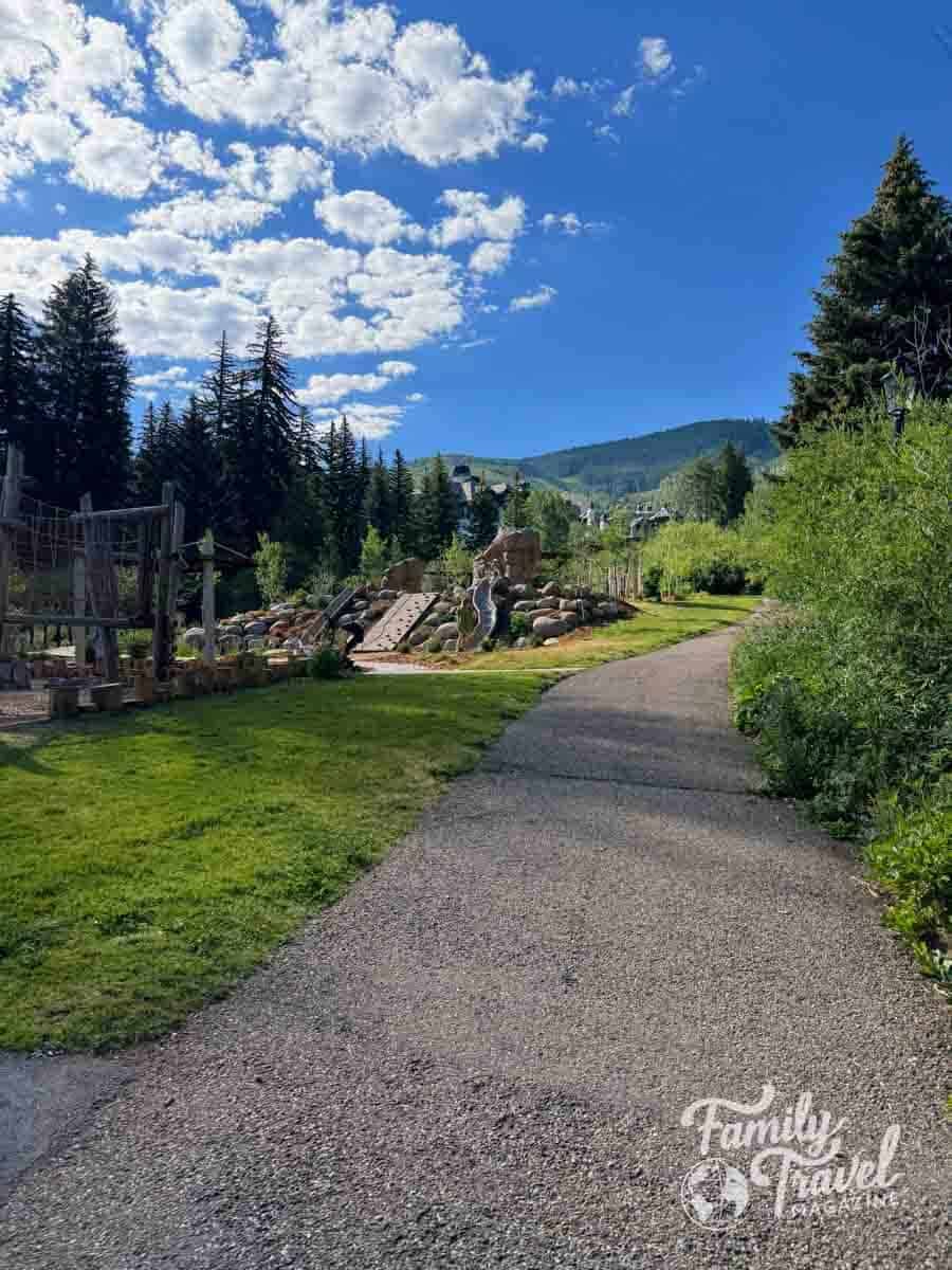 A paved path leads to a natural playground with mountains and resort buildings in Vail, Colorado.