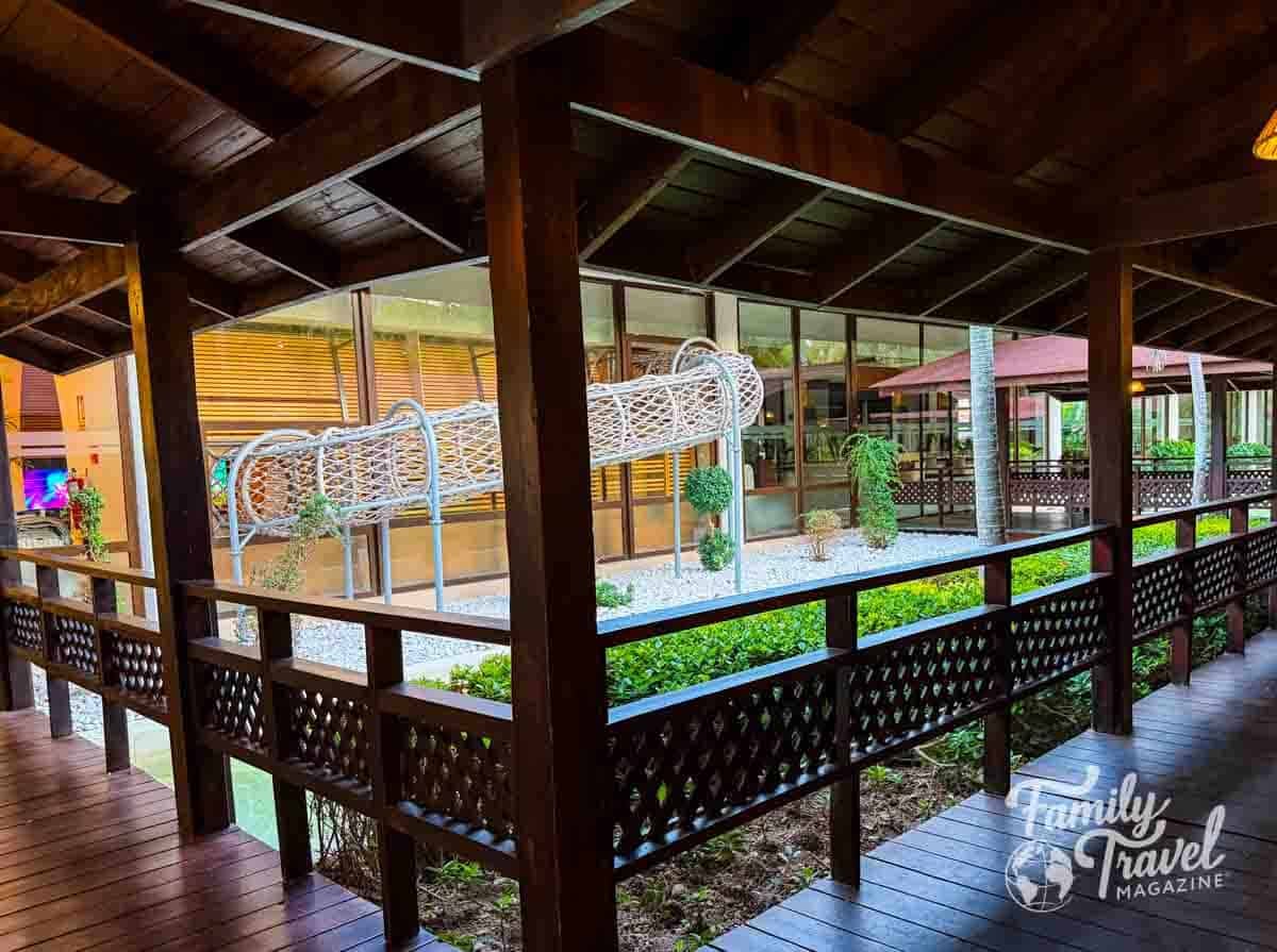 Wooden resort walkway overlooking a courtyard with a white woven play tunnel and lush greenery.
