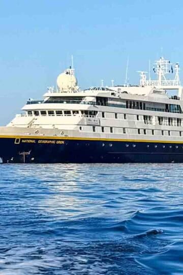 The National Geographic Orion ship with a dark blue hull and white superstructure floats on calm blue water under a clear sky.