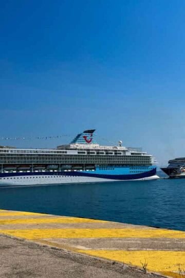 Two large cruise ships are docked at a concrete pier under a clear blue sky.
