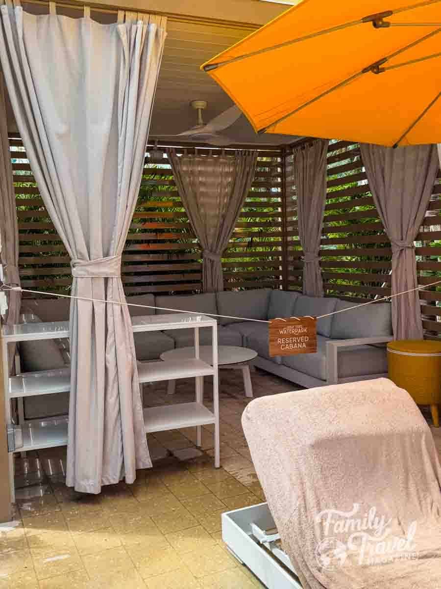 A cabana with gray curtains, a sectional sofa, and a lounge chair sits under a bright orange umbrella.