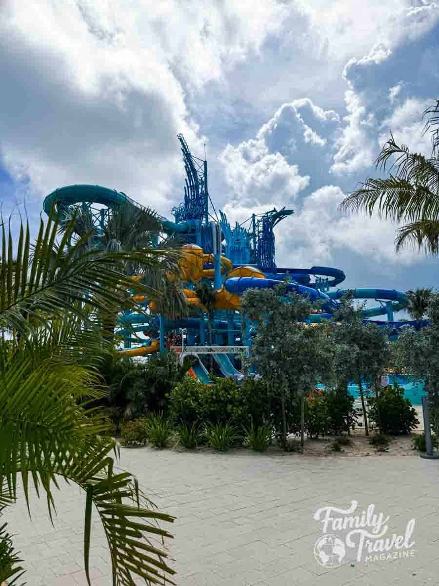 A large, multi-level blue and yellow water slide structure stands behind palm trees under a cloudy sky at Great Tides Waterpark
