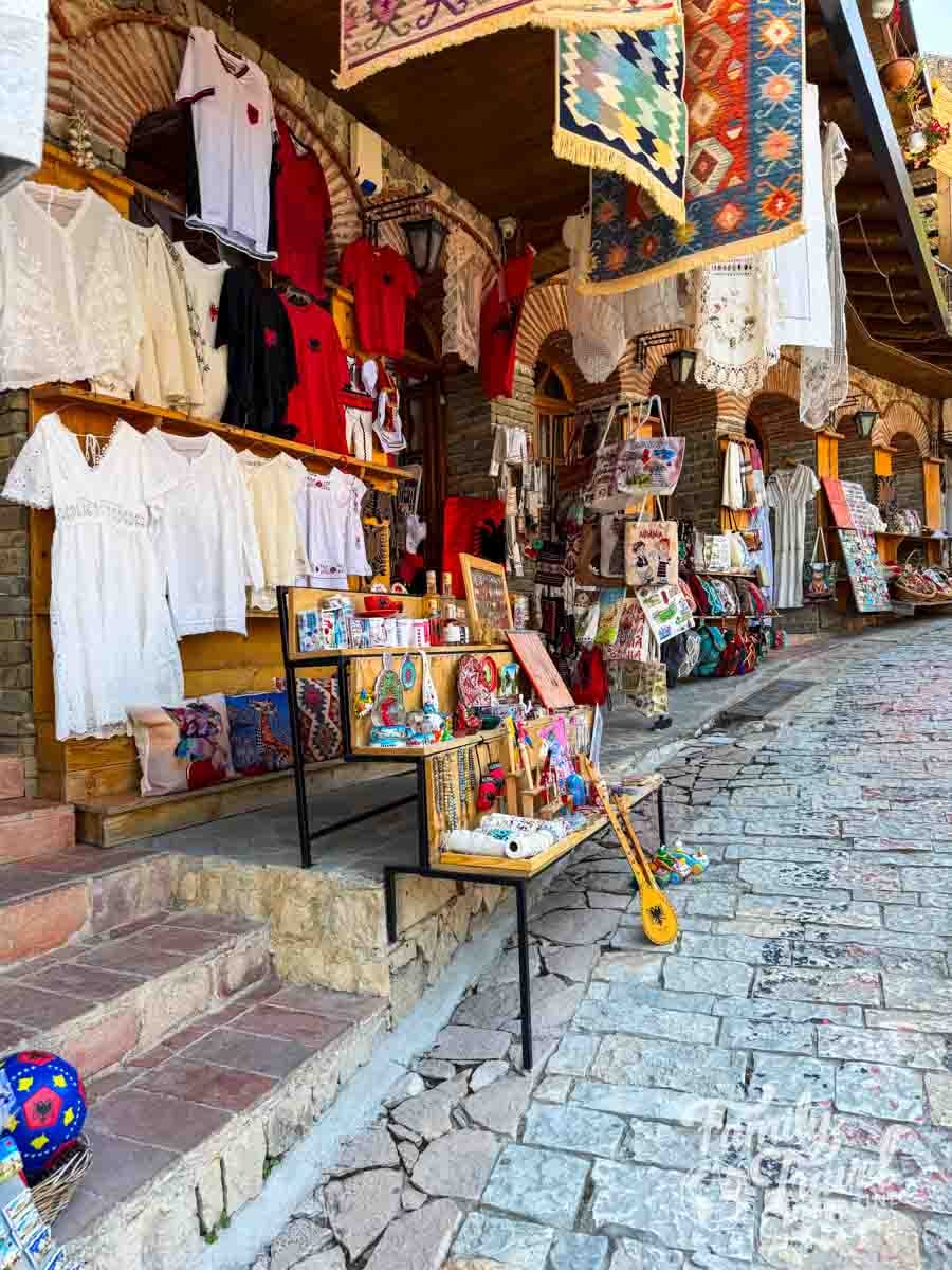 A stone-paved street leads past a shop displaying hanging textiles, clothing, and various souvenirs on wooden shelves.