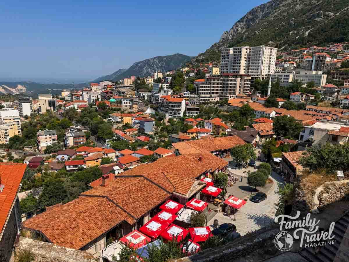A high-angle view shows a town with terracotta-roofed buildings nestled against a mountain slope under a clear blue sky.