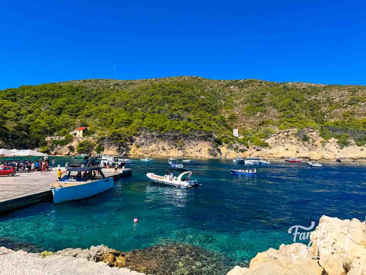 Small boats are docked at a concrete pier in a clear blue bay surrounded by a green, hilly coastline at Bisevo Island. 