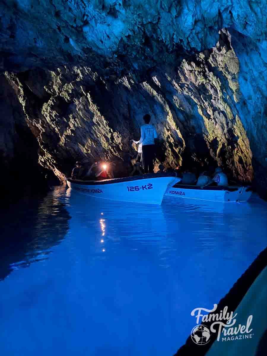 Small boats carrying people float on bright blue water inside a dimly lit cave with rocky walls.