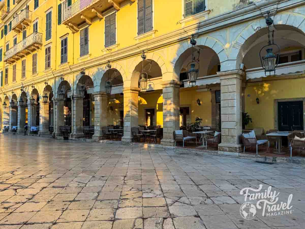 A yellow building with arched walkways and outdoor seating lines a stone-paved plaza in Corfu. 