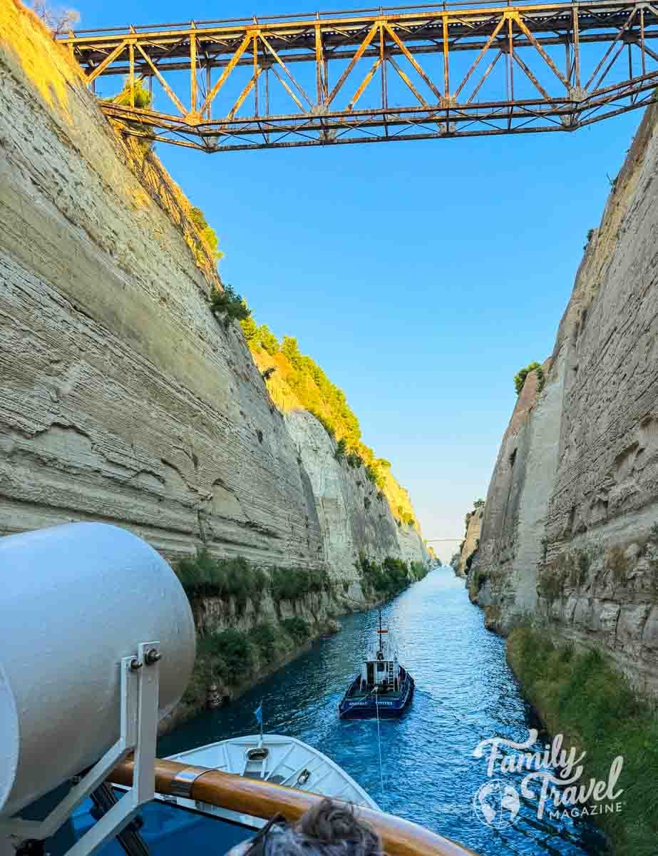 A small boat travels through the narrow Corinth canal between high, steep rock walls under a metal bridge.